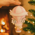 Load image into Gallery viewer, Personalized baby first Christmas ornament featuring a soft pastel pink hot air balloon design with a sweet baby character inside the basket. Handwritten baby name and year or Baby’s First Christmas text displayed on the balloon. Smooth resin finish with gentle glitter accents, nursery themed colors, lightweight structure, mostly 2D ornament with raised 3D details on the front only, designed for baby Christmas tree decor and keepsake gifting.