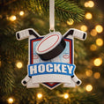Load image into Gallery viewer, Personalized hockey Christmas ornament featuring a hockey puck and crossed hockey sticks design, customizable name area on the front banner and customizable year area on the puck, glossy finish with raised 3D front details and flat 2D back, white hanging ribbon, displayed on a snowy ice rink themed holiday background with hockey accessories and festive winter decorations.
