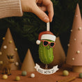Load image into Gallery viewer, Decorative pickle ornament with a Santa hat and sunglasses, held by a hand against a festive background.