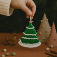 Load image into Gallery viewer, Hand holding a green Christmas tree ornament with a star on top, surrounded by festive decorations.