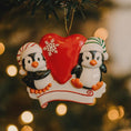 Load image into Gallery viewer, Decorative Christmas ornament with two penguins holding a red heart against a blurred Christmas tree background.