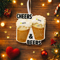 Load image into Gallery viewer, Personalized Cheers and Beers Christmas ornament featuring a raised 3D front with two frothy beer mugs clinking together. The design includes bold âCheersâ and âBeersâ lettering, detailed foam texture, and warm amber beer tones. The ornament has a flat 2D back for easy hanging. Hand-added personalization allows two names and a year. Styled on a rustic wooden background with Christmas lights, greenery, and festive decor for a cozy holiday atmosphere.