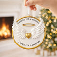 Load image into Gallery viewer, Decorative angel ornament with wings and a heart, held by a hand with a blurred Christmas tree and fireplace in the background.
