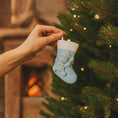 Load image into Gallery viewer, A glossy resin baby boy stocking ornament features a soft pastel blue Christmas stocking shape with a raised glitter style baby figure on the front, white cuff detail, small star accents, and smooth painted surfaces, surrounded by warm holiday lights and nursery themed props, handwritten personalization showing one baby name on the top cuff and the year written near the bottom of the stocking, designed as a mostly 2D ornament with raised 3D details on the front only.