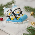 Load image into Gallery viewer, Two penguin figurines on a blue sled with Christmas decorations in the background.