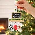 Load image into Gallery viewer, Hand holding a Christmas ornament with stockings and 'We're expecting' sign in front of a decorated tree.