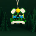 Load image into Gallery viewer, Personalized pickleball Christmas ornament featuring crossed pickleball paddles and a yellow pickleball on a shield style background, customizable name area on the front banner and customizable year area on the ball, glossy finish with raised 3D front details and flat 2D back, white hanging ribbon, displayed on a snowy pickleball court themed holiday background with pine branches and festive decorations.