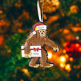 Load image into Gallery viewer, This playful Bigfoot Christmas ornament features a cheerful sasquatch character wearing a Santa hat and holding a gift, finished with a glossy surface that highlights the raised details and festive colors. The design brings a fun and whimsical holiday vibe, making it a standout decoration for Christmas trees, shelves, or seasonal displays, especially for Bigfoot lovers and collectors of unique holiday ornaments.