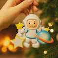 Load image into Gallery viewer, Personalized astronaut Christmas ornament featuring a detailed 3D front design with a smiling space explorer holding a colorful planet and bright yellow stars. The childâs name is added on the suit and the year is written on the star for a custom touch. This space themed ornament is perfect for kids who love rockets, planets, and outer space dreams. A fun and meaningful holiday keepsake for future astronauts and little space lovers.