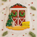 Load image into Gallery viewer, Decorative Christmas scene with a red barn door, wreaths, a tree, and a jar on a snowy background.
