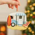 Load image into Gallery viewer, Decorative ornament shaped like a camper with a surfboard and beach chair, held against a blurred Christmas tree background.
