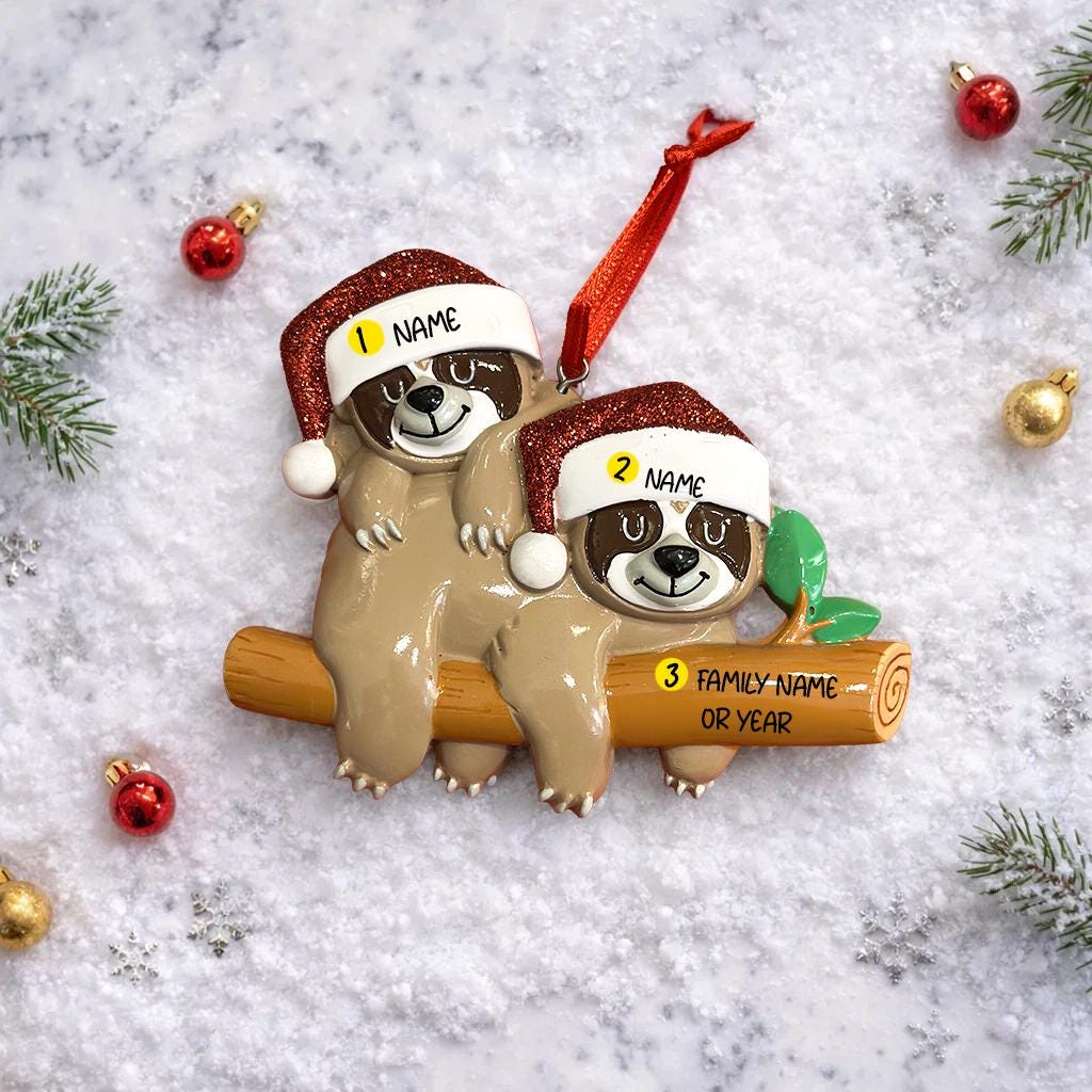 Christmas ornament with two sloths on a log, each wearing a Santa hat, on a snowy background.