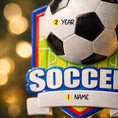 Load image into Gallery viewer, Personalized soccer Christmas ornament featuring a classic black and white soccer ball on a shield style background, customizable name area on the front banner and customizable year area on the soccer ball, glossy finish with raised 3D front details and flat 2D back, white hanging ribbon, displayed on a snowy soccer field themed holiday background with pine branches pinecones and festive decorations.
