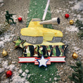 Load image into Gallery viewer, A resin Army tank ornament in bold yellow features a cartoon style military tank with detailed black tracks and raised cannon, camouflage green ARMY lettering across the front, a star accent and American flag inspired base, surrounded by snow texture and festive holiday elements, with handwritten personalization showing one name on the left banner and the year on the right banner, designed as a mostly 2D ornament with raised 3D details on the front only for lightweight Christmas tree decor.
