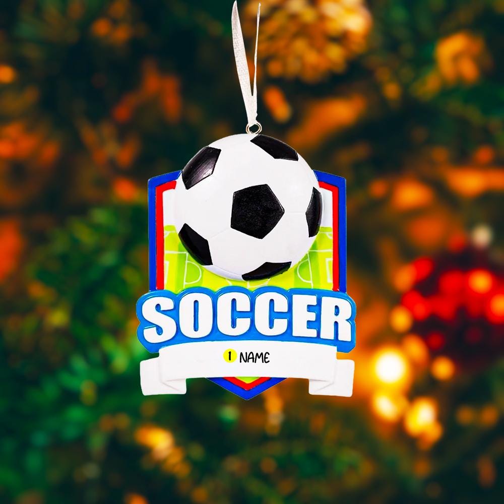 Personalized soccer Christmas ornament featuring a classic black and white soccer ball on a shield style background, customizable name area on the front banner and customizable year area on the soccer ball, glossy finish with raised 3D front details and flat 2D back, white hanging ribbon, displayed on a snowy soccer field themed holiday background with pine branches pinecones and festive decorations.