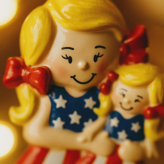 Decorative figurine of a girl holding a smaller figure, both wearing red, white, and blue outfits.