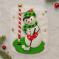Load image into Gallery viewer, Decorative snowman ornament with a golf club and flag on a snowy background
