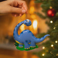 Load image into Gallery viewer, Blue dinosaur ornament held by a hand with a Christmas tree in the background
