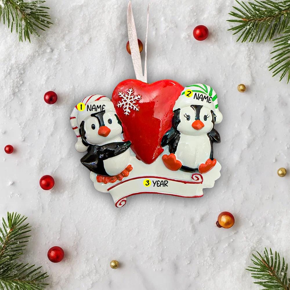 Personalized Christmas ornament with two penguins holding a heart, surrounded by festive decorations on a snowy background.