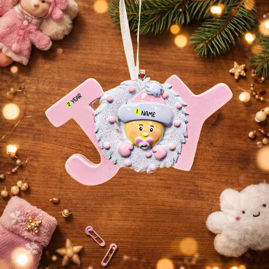 Decorative ornament shaped like a baby in a pink outfit with a name tag, surrounded by Christmas decorations on a wooden surface.