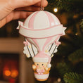 Load image into Gallery viewer, Personalized baby first Christmas ornament featuring a soft pastel pink hot air balloon design with a sweet baby character inside the basket. Handwritten baby name and year or Baby’s First Christmas text displayed on the balloon. Smooth resin finish with gentle glitter accents, nursery themed colors, lightweight structure, mostly 2D ornament with raised 3D details on the front only, designed for baby Christmas tree decor and keepsake gifting.
