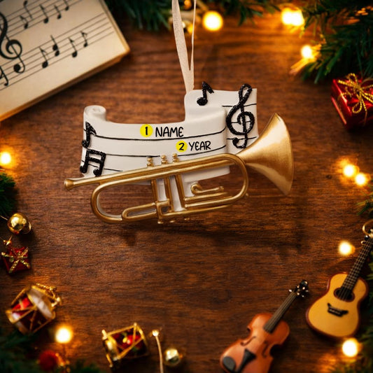 Personalized music ornament featuring a white musical staff design with black notes and a gold trumpet detail. Includes free handwritten personalization with one name and year written on the sheet music. Shown hanging with a ribbon in a warm festive holiday setting with soft lights instruments and seasonal decor. Designed with a 3D raised front and flat back perfect for Christmas tree display and gifts for musicians music lovers and band members.