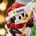 Load image into Gallery viewer, Penguin ornament with a Santa hat and name tag, holding a yellow stick, against a blurred Christmas tree background.
