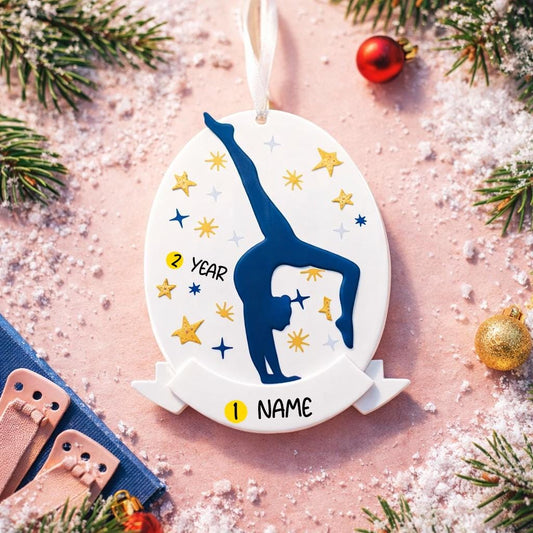 Personalized gymnastics Christmas ornament featuring an oval white design with a navy blue gymnast silhouette performing a handstand pose, surrounded by gold and blue star accents, customizable year displayed on the upper area, customizable name space on the bottom banner, smooth glossy finish with raised 3D front details and flat 2D back, white hanging ribbon, photographed on a soft pink snowy holiday background with winter greenery, festive ornaments, and warm seasonal styling.
