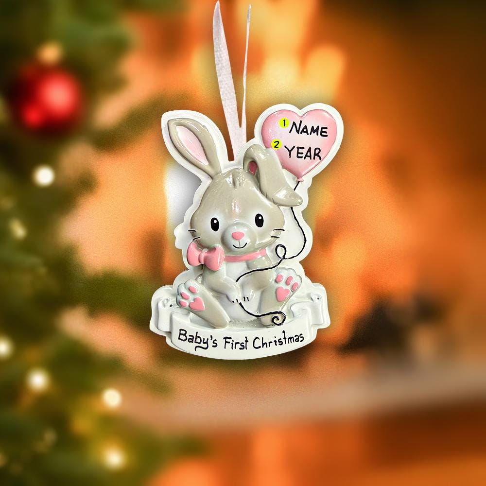 Decorative Christmas ornament of a baby rabbit with a heart-shaped name tag against a blurred Christmas tree background.