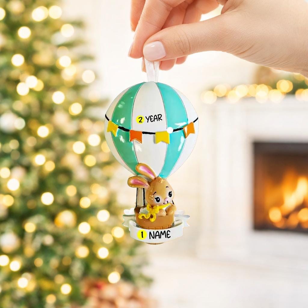 A personalized baby boy ornament featuring a cute bunny riding a pastel blue hot air balloon. Soft blue and white tones with gentle decorative details create a calm and cozy look. Handwritten personalization includes baby name and year. Mostly 2D ornament with raised 3D details on the front only, lightweight and easy to hang, perfect for baby first Christmas, nursery decor, and toddler keepsakes.