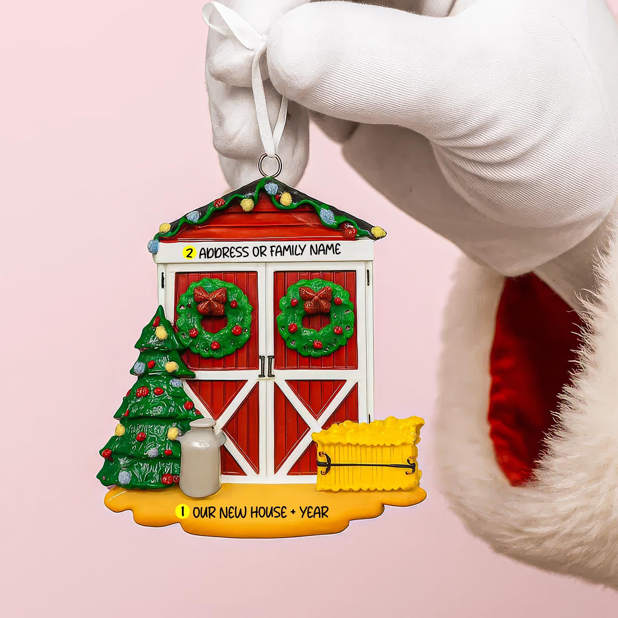 Decorative Christmas ornament shaped like a house with festive elements, held by a white glove on a pink background.