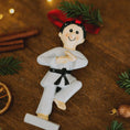 Load image into Gallery viewer, Decorative ornament of a martial artist in a white gi with a black belt on a wooden surface with Christmas decorations.
