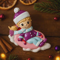 Load image into Gallery viewer, Christmas ornament of a girl in a pink sleigh with purple hat and coat on a wooden background.

