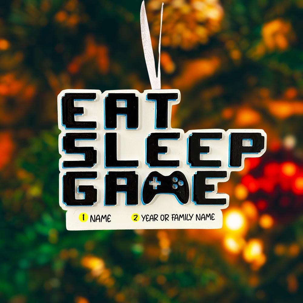 Personalized Eat Sleep Game Christmas ornament with a raised 3D front featuring bold block-style lettering and a detailed game controller icon. The front design is fully three dimensional, creating depth and texture, while the back is flat 2D for easy hanging and durability. White base with black raised letters and subtle colored accents. Includes hand-added personalization with name and year or family name. Gaming-themed holiday ornament displayed on snowy festive background with warm lights, designed for 