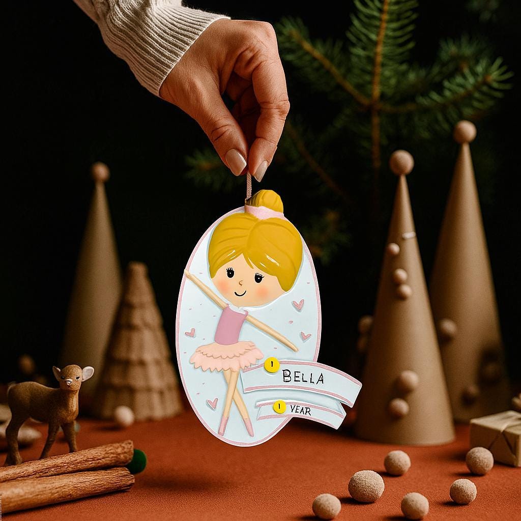 Personalized ballet girl Christmas ornament featuring a cute ballerina character in a pink tutu dress with heart details, customizable name area on the front banner and customizable year area below, glossy finish with raised 3D front details and flat 2D back, white hanging ribbon, displayed on a snowy holiday background with pine branches and festive decorations.