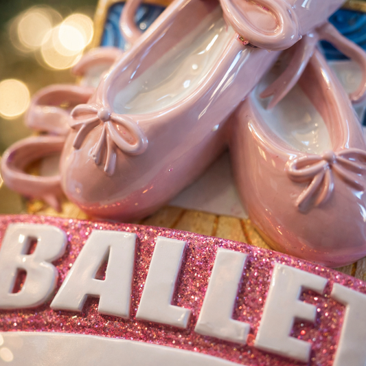 Personalized ballet Christmas ornament featuring soft pink ballet shoes with delicate ribbon details and a glittered ballet banner, customizable name area on the lower front ribbon and customizable year area beside the name, glossy finish with raised 3D front elements and flat 2D back, white hanging ribbon, displayed on a warm snowy holiday background with festive greenery ornaments and cozy Christmas decor.