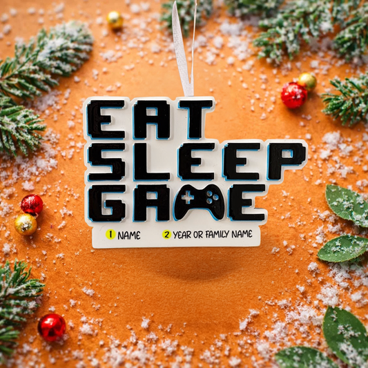 Personalized Eat Sleep Game Christmas ornament with a raised 3D front featuring bold block-style lettering and a detailed game controller icon. The front design is fully three dimensional, creating depth and texture, while the back is flat 2D for easy hanging and durability. White base with black raised letters and subtle colored accents. Includes hand-added personalization with name and year or family name. Gaming-themed holiday ornament displayed on snowy festive background with warm lights, designed for 