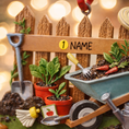 Load image into Gallery viewer, Personalized gardener ornament featuring a detailed garden scene with wooden fence wheelbarrow potted plants gardening tools and soil elements. The design includes a customizable name on the fence and a year on the base making it a thoughtful keepsake for garden lovers. The ornament has a warm rustic farmhouse style and is displayed with a red hanging ribbon suitable for Christmas trees seasonal decor and gift displays.
