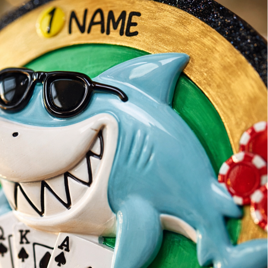 Personalized poker ornament featuring a cool shark character wearing sunglasses with playing cards and red casino chips. Designed in a round ornament style with a glossy finish. Includes free handwritten personalization with one name and year displayed on the front. Shown hanging with a ribbon in a festive holiday setting with greenery snow and seasonal decor. 3D raised front and flat back design perfect for Christmas trees and gifts for poker players casino lovers and card game fans.