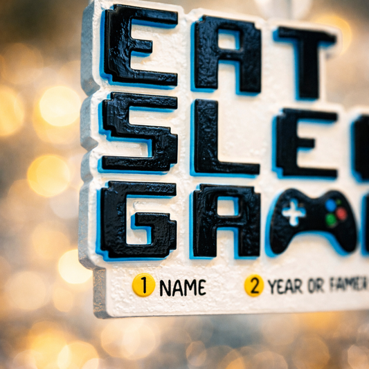 Personalized Eat Sleep Game Christmas ornament with a raised 3D front featuring bold block-style lettering and a detailed game controller icon. The front design is fully three dimensional, creating depth and texture, while the back is flat 2D for easy hanging and durability. White base with black raised letters and subtle colored accents. Includes hand-added personalization with name and year or family name. Gaming-themed holiday ornament displayed on snowy festive background with warm lights, designed for 