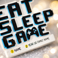 Load image into Gallery viewer, Personalized Eat Sleep Game Christmas ornament with a raised 3D front featuring bold block-style lettering and a detailed game controller icon. The front design is fully three dimensional, creating depth and texture, while the back is flat 2D for easy hanging and durability. White base with black raised letters and subtle colored accents. Includes hand-added personalization with name and year or family name. Gaming-themed holiday ornament displayed on snowy festive background with warm lights, designed for 
