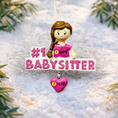 Load image into Gallery viewer, Personalized Babysitter Christmas ornament featuring a detailed raised 3D front design with glitter pink lettering and a charming girl figure. Custom name is added on the shirt and year is written on the hanging heart charm. Bright pink and white holiday colors stand out beautifully on the tree. Flat 2D back for easy hanging. Perfect keepsake gift for babysitter appreciation, nanny thank you present, childcare provider Christmas decor.
