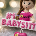 Load image into Gallery viewer, Personalized Babysitter Christmas ornament featuring a detailed raised 3D front design with glitter pink lettering and a charming girl figure. Custom name is added on the shirt and year is written on the hanging heart charm. Bright pink and white holiday colors stand out beautifully on the tree. Flat 2D back for easy hanging. Perfect keepsake gift for babysitter appreciation, nanny thank you present, childcare provider Christmas decor.
