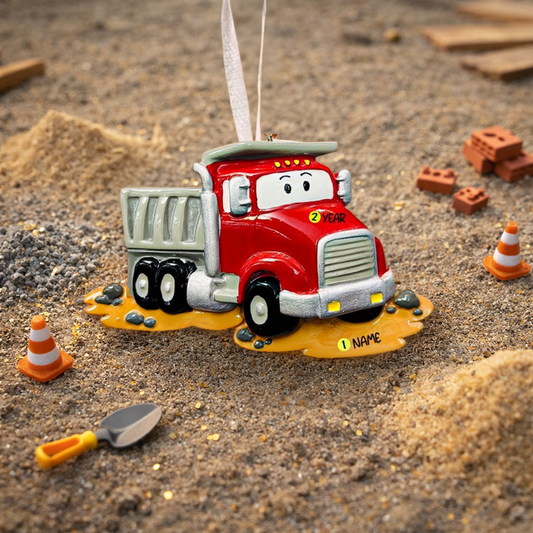 Red toy dump truck ornament on a sandy surface with construction-themed decorations.
