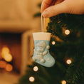 Load image into Gallery viewer, A glossy resin baby boy stocking ornament features a soft pastel blue Christmas stocking shape with a raised glitter style baby figure on the front, white cuff detail, small star accents, and smooth painted surfaces, surrounded by warm holiday lights and nursery themed props, handwritten personalization showing one baby name on the top cuff and the year written near the bottom of the stocking, designed as a mostly 2D ornament with raised 3D details on the front only.
