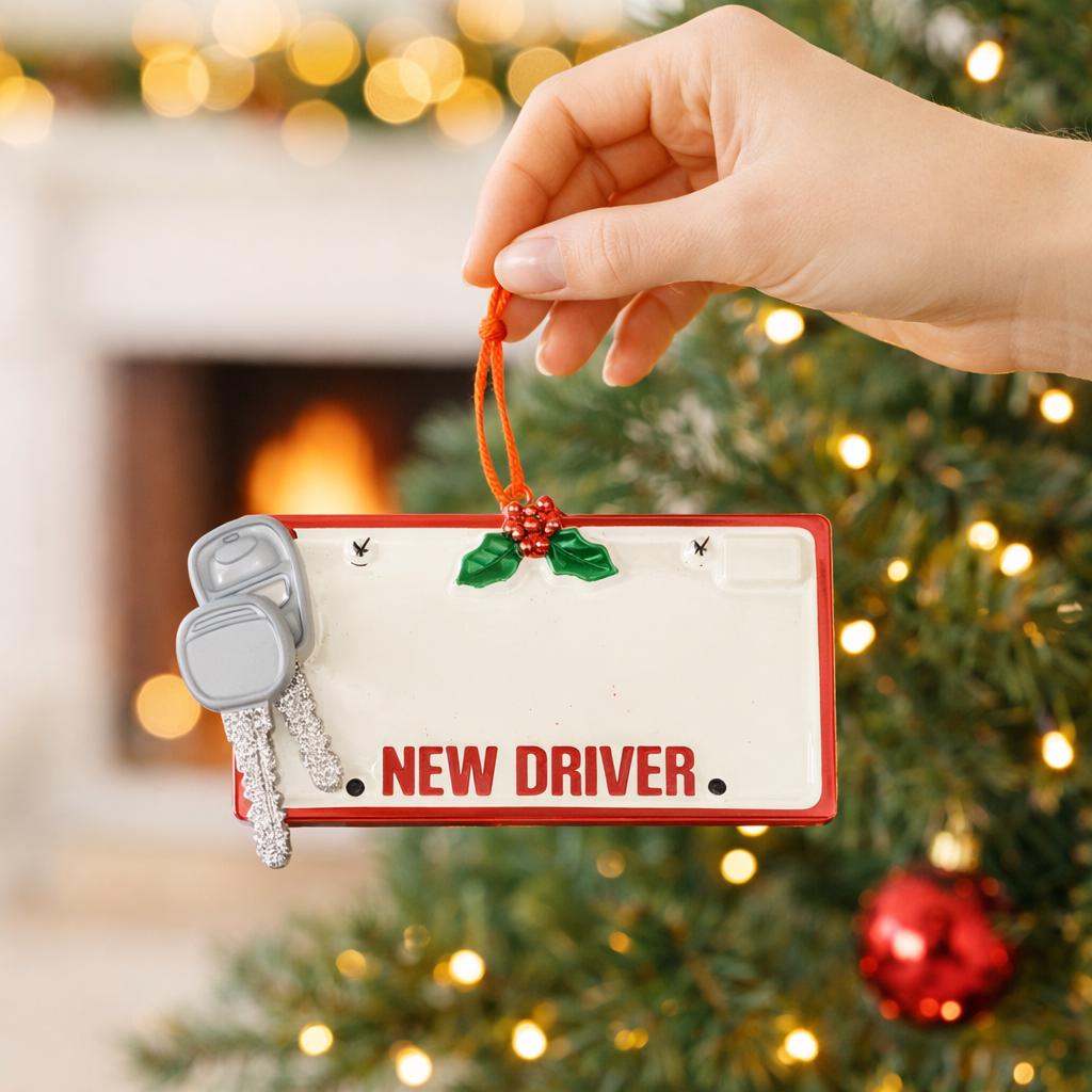 Hand holding a Christmas ornament with a 'New Driver' sign and car keys in front of a decorated tree.