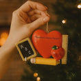 Load image into Gallery viewer, Decorative ornament with heart, apple, and chalkboard held by a hand against a blurred Christmas tree background.
