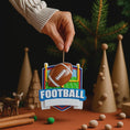 Load image into Gallery viewer, Personalized football Christmas ornament featuring a brown football with stitched details on a shield style background and goalpost design, customizable name area on the front banner and customizable year area on the football, glossy finish with raised 3D front details and flat 2D back, white hanging ribbon, displayed on a snowy football field themed holiday background with pine branches and festive decorations.
