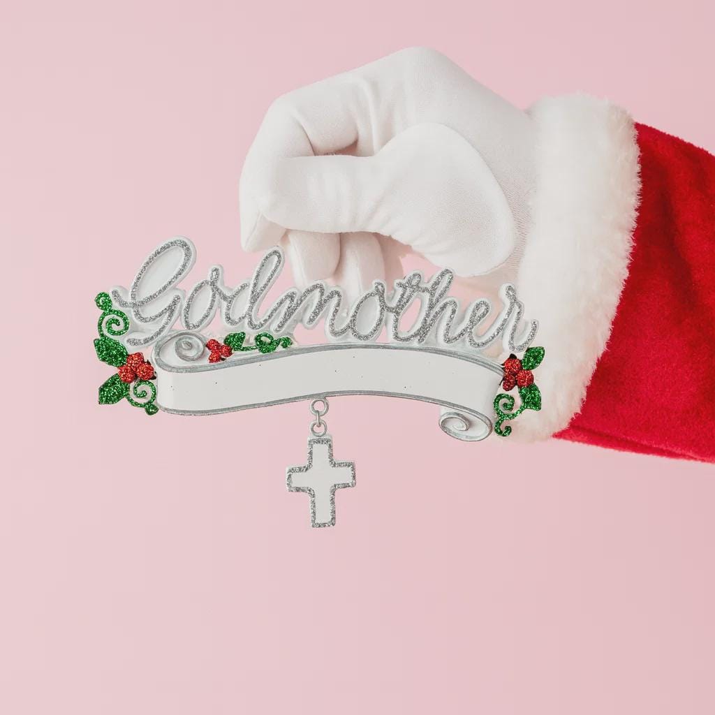 Decorative ornament with 'Godmother' text held by a Santa Claus glove on a pink background