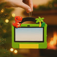 Load image into Gallery viewer, Decorative ornament with a dinosaur and palm tree on a green frame, held against a festive background.
